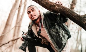 Man ducking under a tree trunk with a camera around his neck
